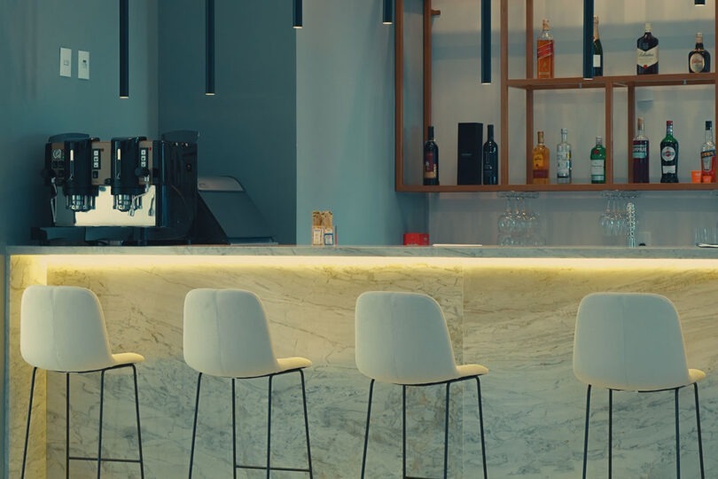 Bar do CLATT Hotel com cafeteira profissional, estante de design contemporâneo com garrafas de bebidas para o preparo de drinks, balcão com cadeiras altas estofadas