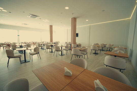 Mesas de madeira com poltronas estofadas do restaurante do CLATT Hotel, com TV na parede e ambiente bem iluminado