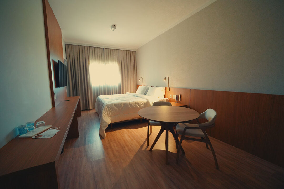 Vista do quarto com cama, enxoval premium, cortinas blackout, mesa redonda com duas cadeiras estofadas, TV, estação de trabalho.