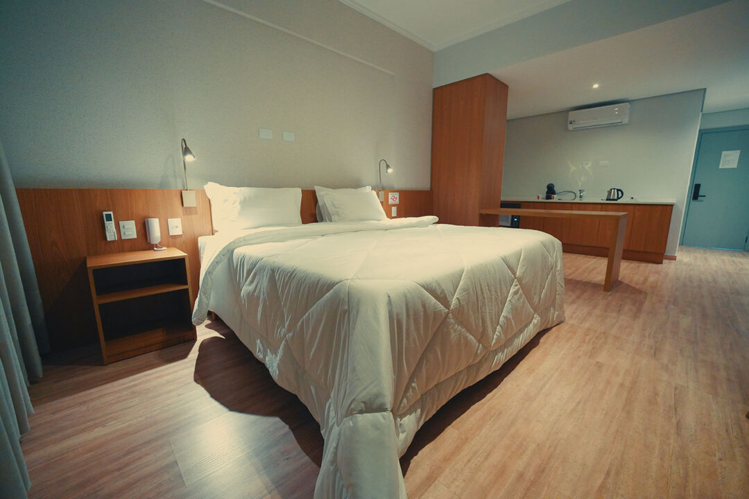 Cama com enxoval premium, cabeceira de madeira, vista parcial da cozinha com forno elétrico, cafeteira, chaleira elétrica, pia e ar condicionado.