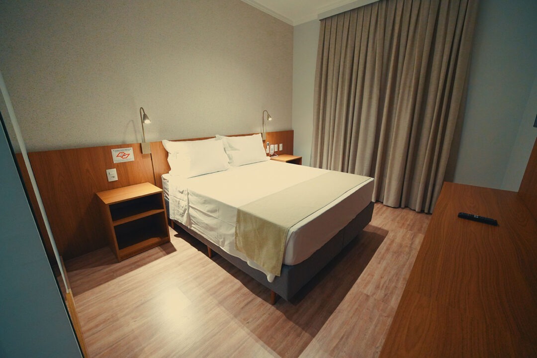 Cama com cabeceira de madeira, enxoval premium, controle do ar condicionado e luminária para leitura, bem como cortinas do chão ao teto.