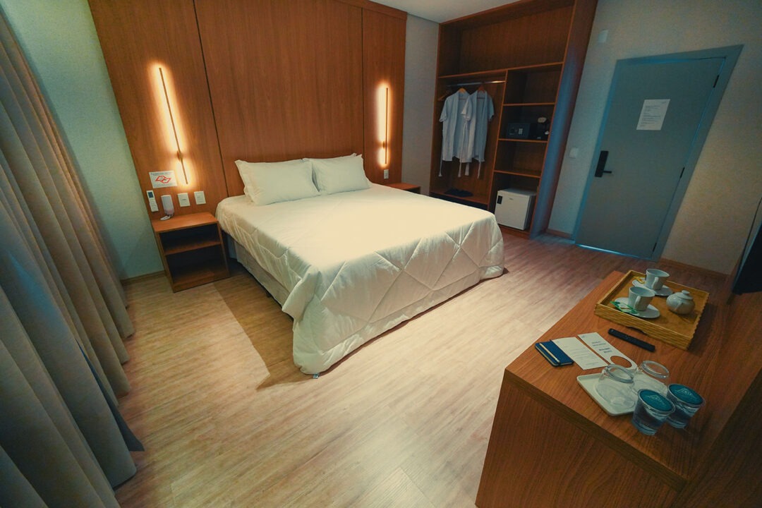 Cama com enxoval premium, guarda roupa com roupão, cofre, frigobar, estação de trabalho, tv, cortinas, iluminação na cabeceira de madeira da cama.