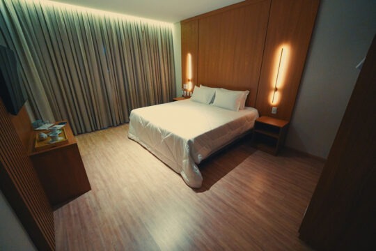 Vista do quarto com cama, enxoval premium, cortinas blackout, TV.