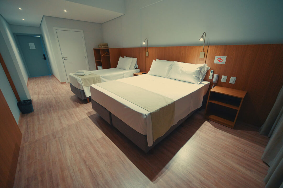 Duas camas com enxoval, cortinas, Telefone na cabeceira junto à luminária para leitura, mesinha de cabeceira e cabeceira de madeira.