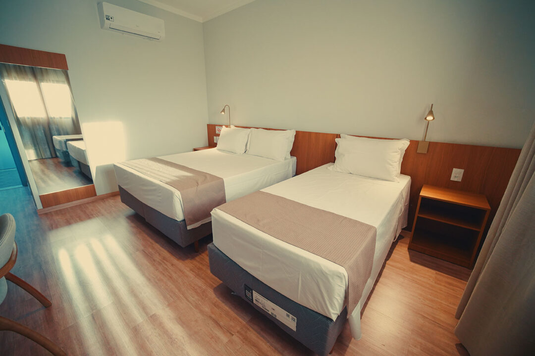 Duas camas com enxoval, cortinas, quarto iluminado com cabeceira de madeira, ar condicionado e luminária para leitura.