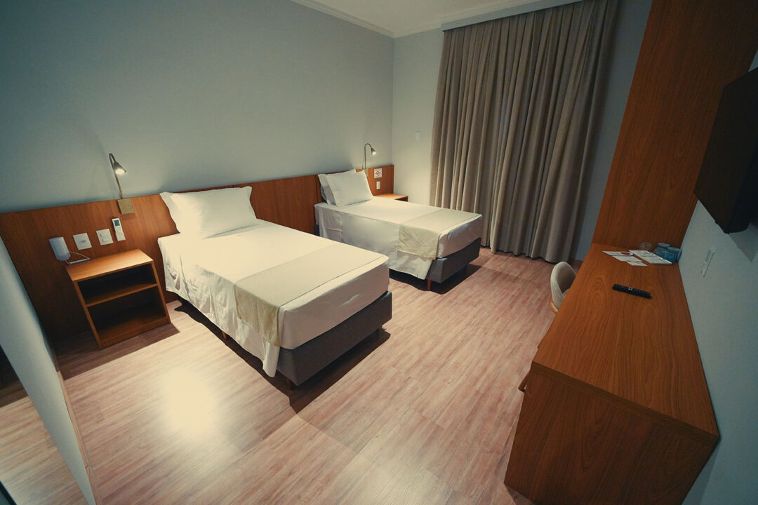 Duas camas com enxoval, cortinas, cabeceira de madeira com mesinha de apoio e luminárias para leitura, além de TV e estação de trabalho.